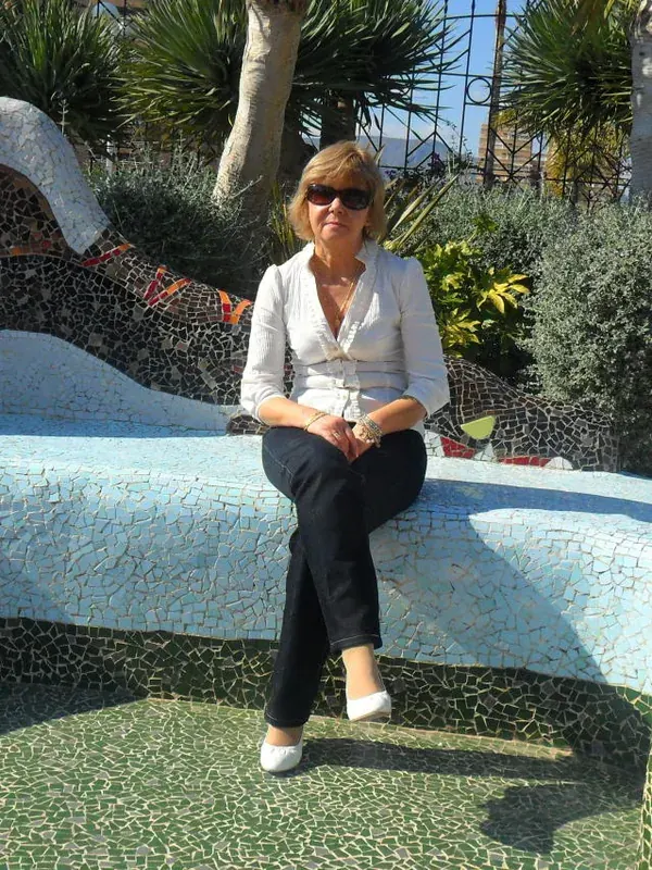Personne assise sur un banc en mosaïque bleue, devant une sculpture d'œil en mosaïque, dans un jardin ensoleillé avec plantes et palmiers.