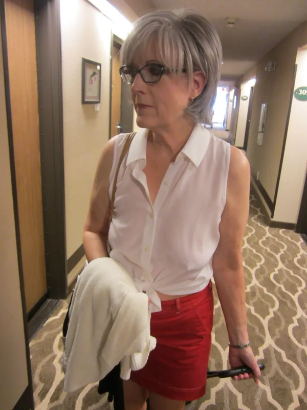 Personne aux cheveux gris portant une chemise blanche et une jupe rouge, tenant une valise dans un couloir d'hôtel