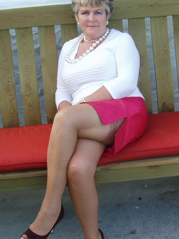 Personne assise sur banc en bois, chemise blanche, jupe rose, chaussures à talons, perles, coussin rouge