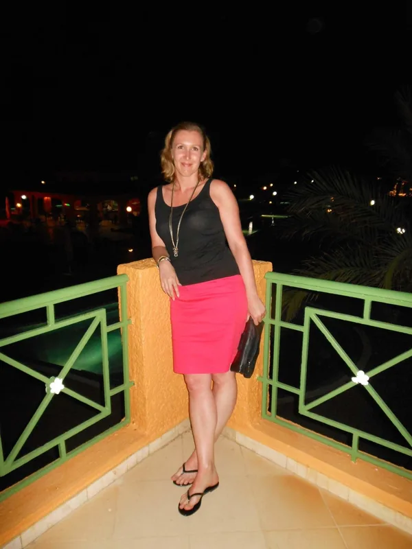 Personne en tenue colorée sur terrasse la nuit, avec jupe rose, haut noir, sandales, rambarde verte et piscine éclairée.