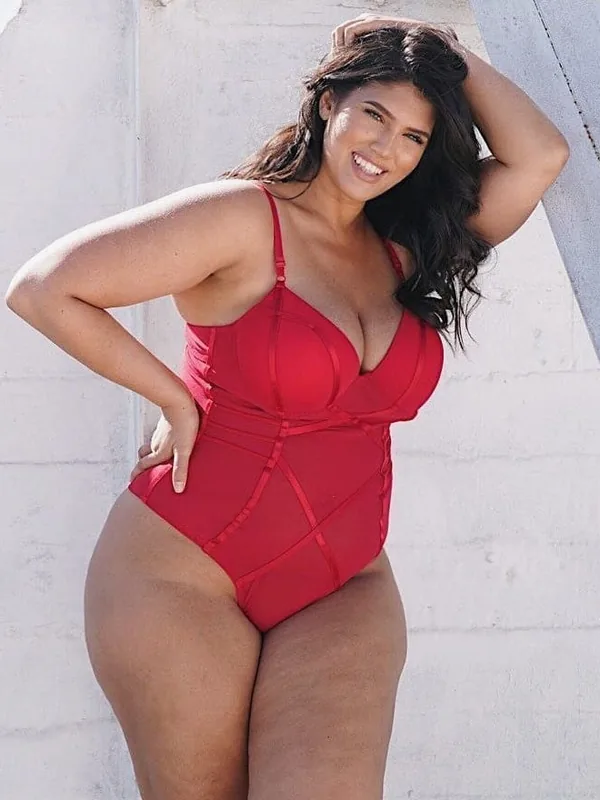 Personne en maillot de bain rouge posant contre un mur blanc, souriante, main dans les cheveux
