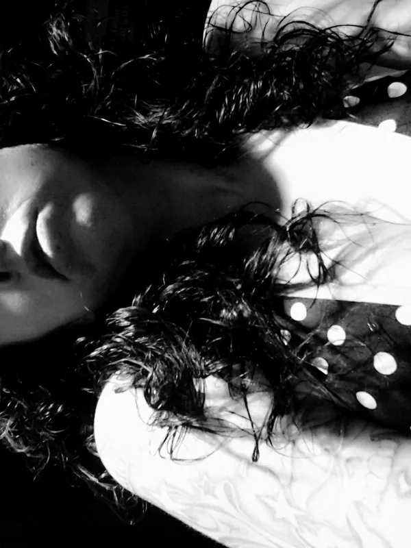 Portrait noir et blanc d'une personne allongée, cheveux bouclés, regard direct, haut à pois, bras tatoué