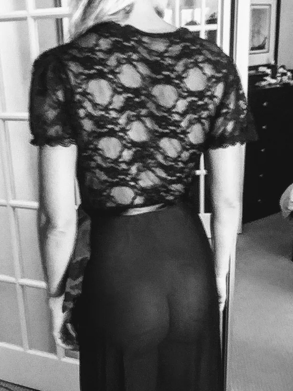 Personne de dos en robe noire à dentelle, cheveux blonds, près d'une porte vitrée, photo en noir et blanc
