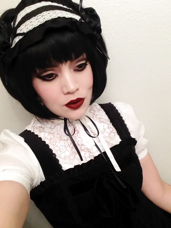 Portrait de profil d'une personne en tenue gothique lolita, cheveux noirs, maquillage sombre, salopette noire et chemise blanche à dentelle.