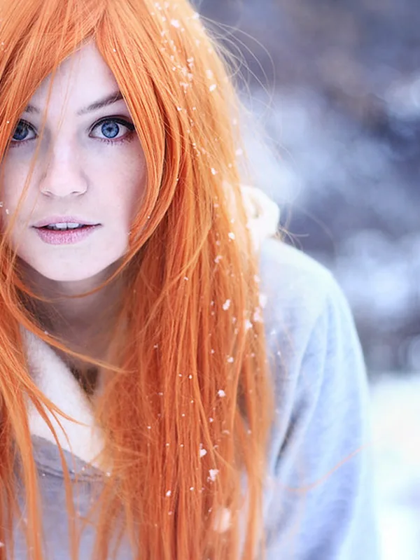 Portrait en hiver d'une personne aux cheveux roux et yeux bleus, sous la neige, vêtue d'un pull gris à capuche.
