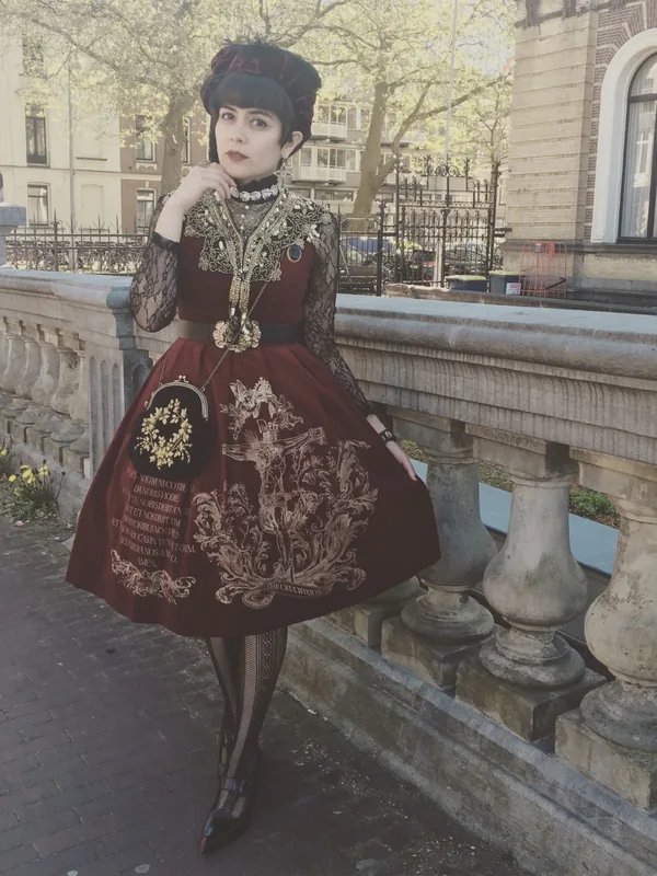 Portrait d'une personne en robe bordeaux ornée de motifs religieux dorés, chapeau sombre, collants à mailles