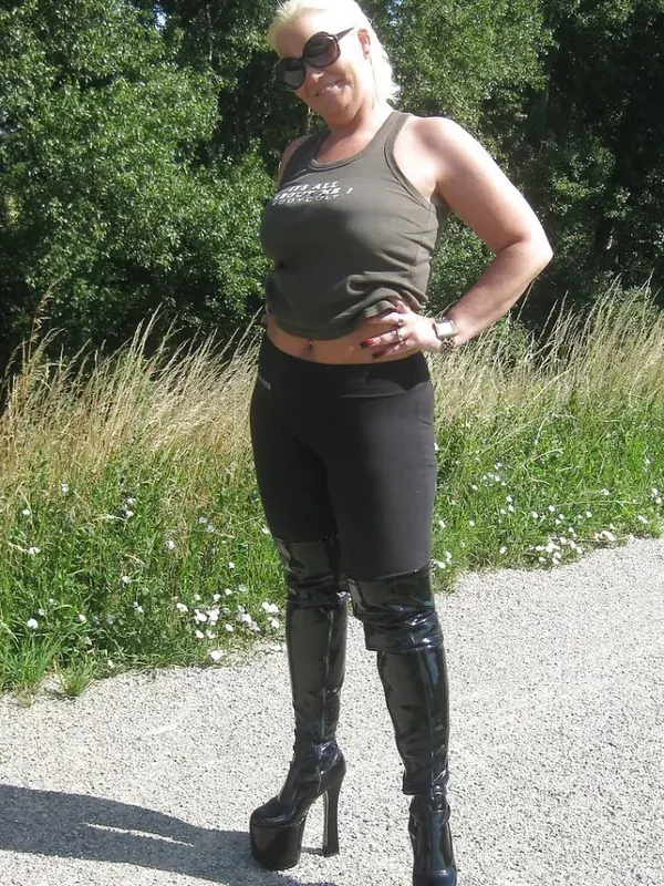 Personne en tenue sombre avec bottes à talons hauts, lunettes de soleil, debout sur un chemin gravillonné entouré de verdure.