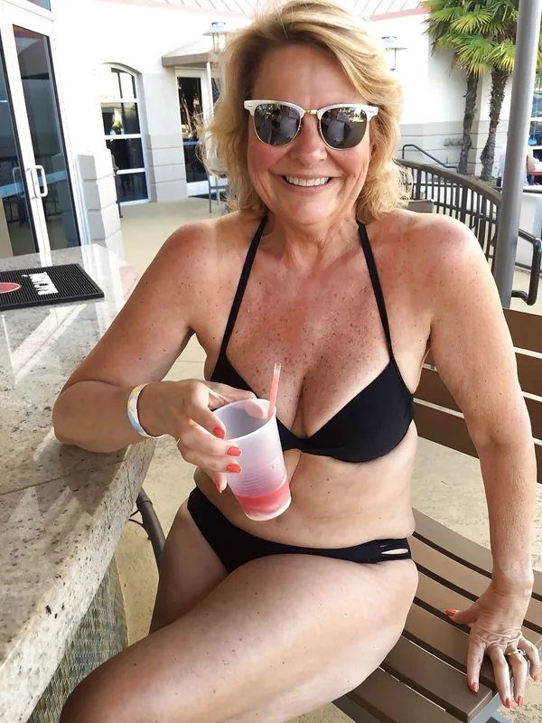 Personne en maillot noir, lunettes de soleil, tenant un gobelet rose, assise à l'extérieur sous le soleil