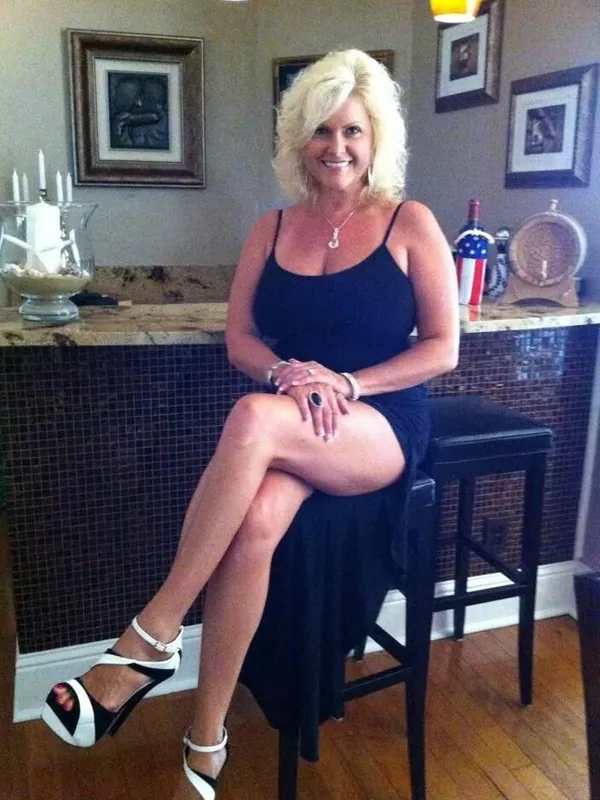 Personne aux cheveux blonds en robe noire, assise sur tabouret, souriante, dans un intérieur chaleureux avec comptoir et décorations.
