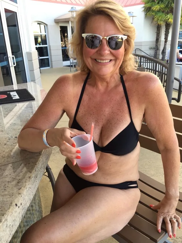 Personne en maillot de bain noir, lunettes de soleil, tenant un gobelet, assise à une table extérieure ensoleillée.