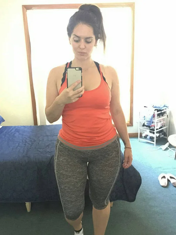 Femme prenant un selfie devant un miroir dans une chambre, vêtue d'un haut orange et de leggings gris.