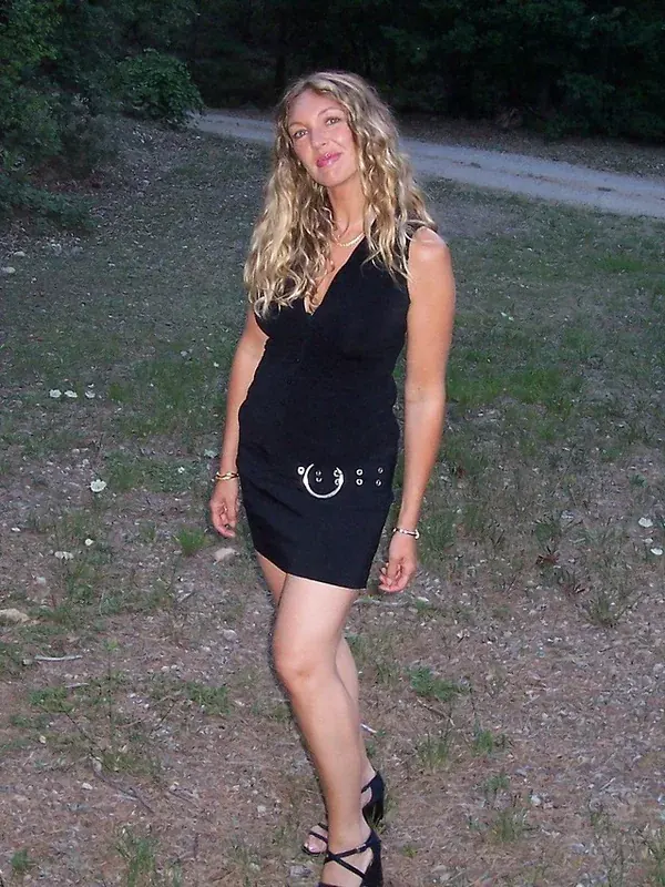Femme aux cheveux blonds portant une robe noire courte, debout sur un terrain herbeux en extérieur.